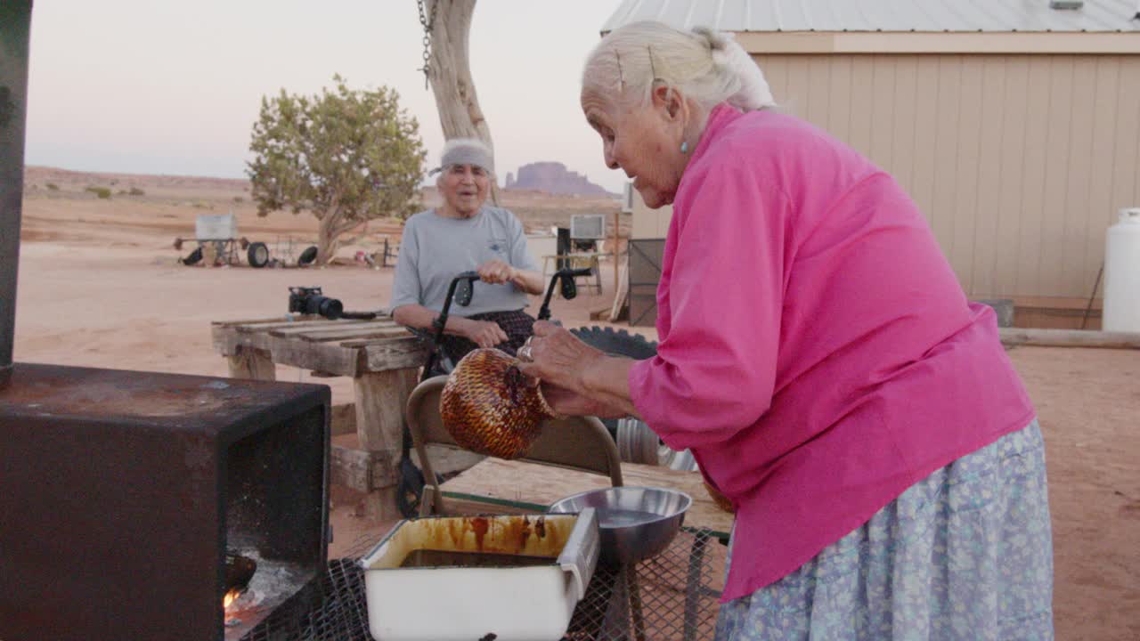 上了年纪的纳瓦霍妇女在她新编织的篮子上涂上松树树沥青，在室外的木炉子上取暖，她的丈夫和孙子看着她工作视频下载