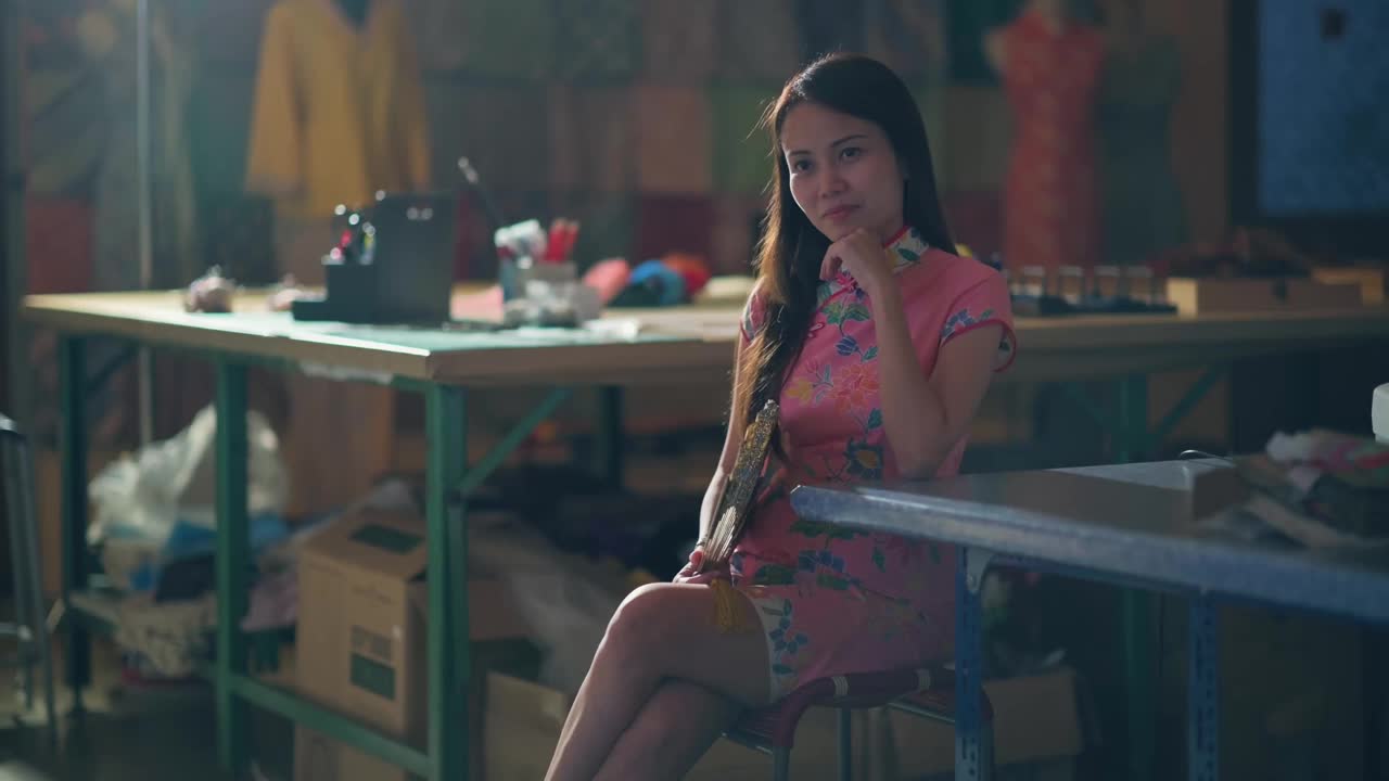 亚洲华裔女裁缝小企业主坐在她的工作地点，穿着中国传统服装旗袍，手拿折扇，下巴上带着自信和满意的微笑视频下载