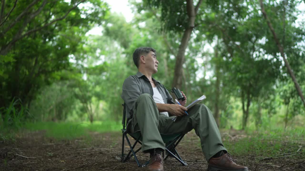 亚洲老人，退休了，坐在椅子上用双筒望远镜看树上的鸟，在树林里，并把信息写在笔记本上。快乐地度过假期视频下载