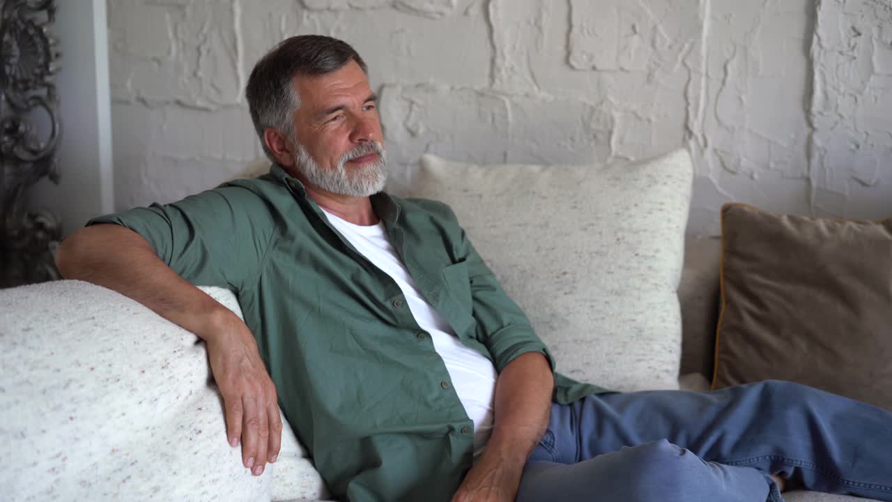 微笑的成熟男人在沙发上放松。白发苍苍的自信老人在家看着摄像机视频素材