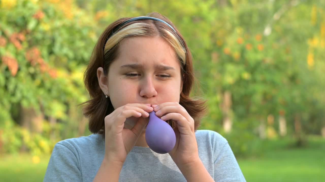 在一个绿色公园里，一个染了头发，染了头发的红发少女正在努力地给一个淡紫色的气球充气，这是一个特写视频肖像。视频下载