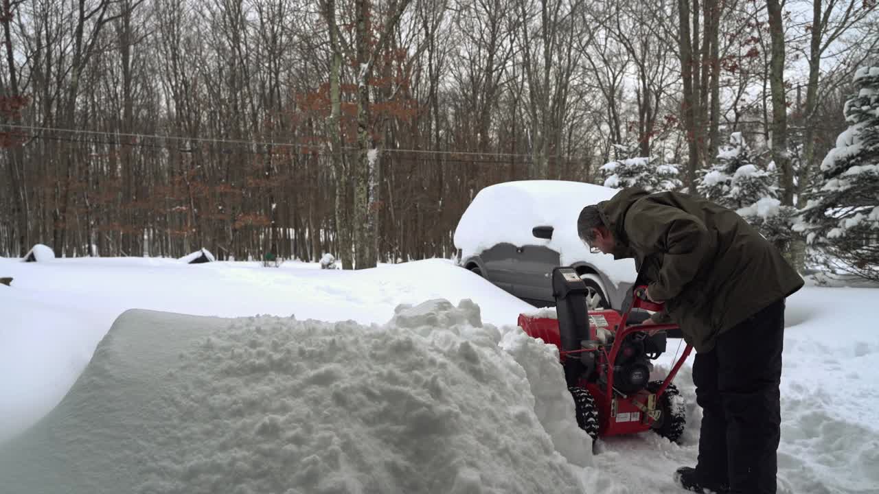 一场冬季暴风雪过后，一名男子正在清理他被雪覆盖的汽车周围的车道。他正在启动除雪机。视频下载
