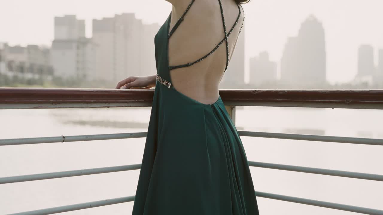 一位身穿绿色晚礼服的年轻亚洲新娘，站在湖滨码头，以城市建筑为背景，微笑着看着镜头，用慢镜头拍摄视频下载