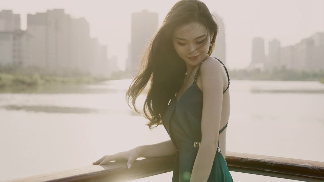 一位身穿绿色晚礼服的年轻亚洲新娘，站在湖滨码头，以城市建筑为背景，微笑着看着镜头，用慢镜头拍摄视频素材