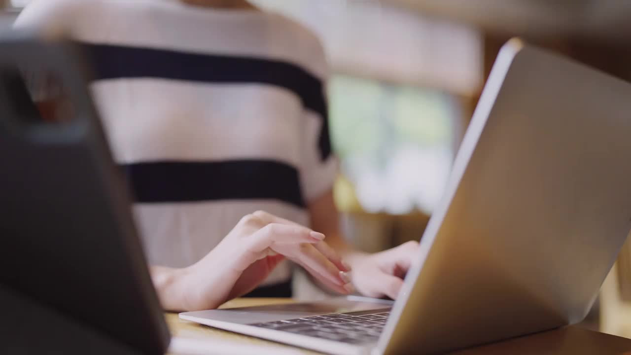 靠近年轻的亚洲女性用户工人的女性手在笔记本电脑上打字，笔记本电脑键盘坐在咖啡馆的桌子上工作，与笔记本电脑软件应用程序技术概念视频素材