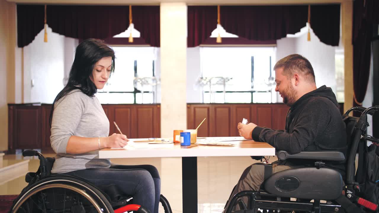 轮椅。残疾。残疾青年男女坐在轮椅上，坐在桌旁用颜料画画。为残疾人提供的创造性团体课程。教育、关系与人文观念视频素材