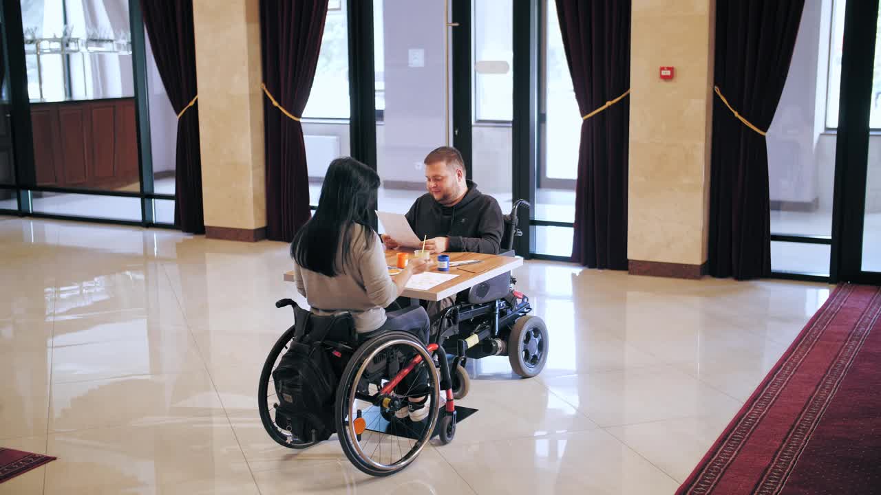 轮椅。残疾。残疾青年男女坐在轮椅上，坐在桌旁用颜料画画。为残疾人提供的创造性团体课程。教育、关系与人文观念视频素材