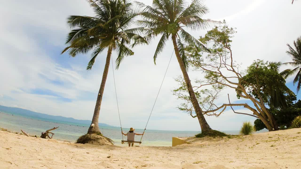 快乐的年轻女子在椰子树上荡秋千，看着令人惊叹的热带海滩视频下载