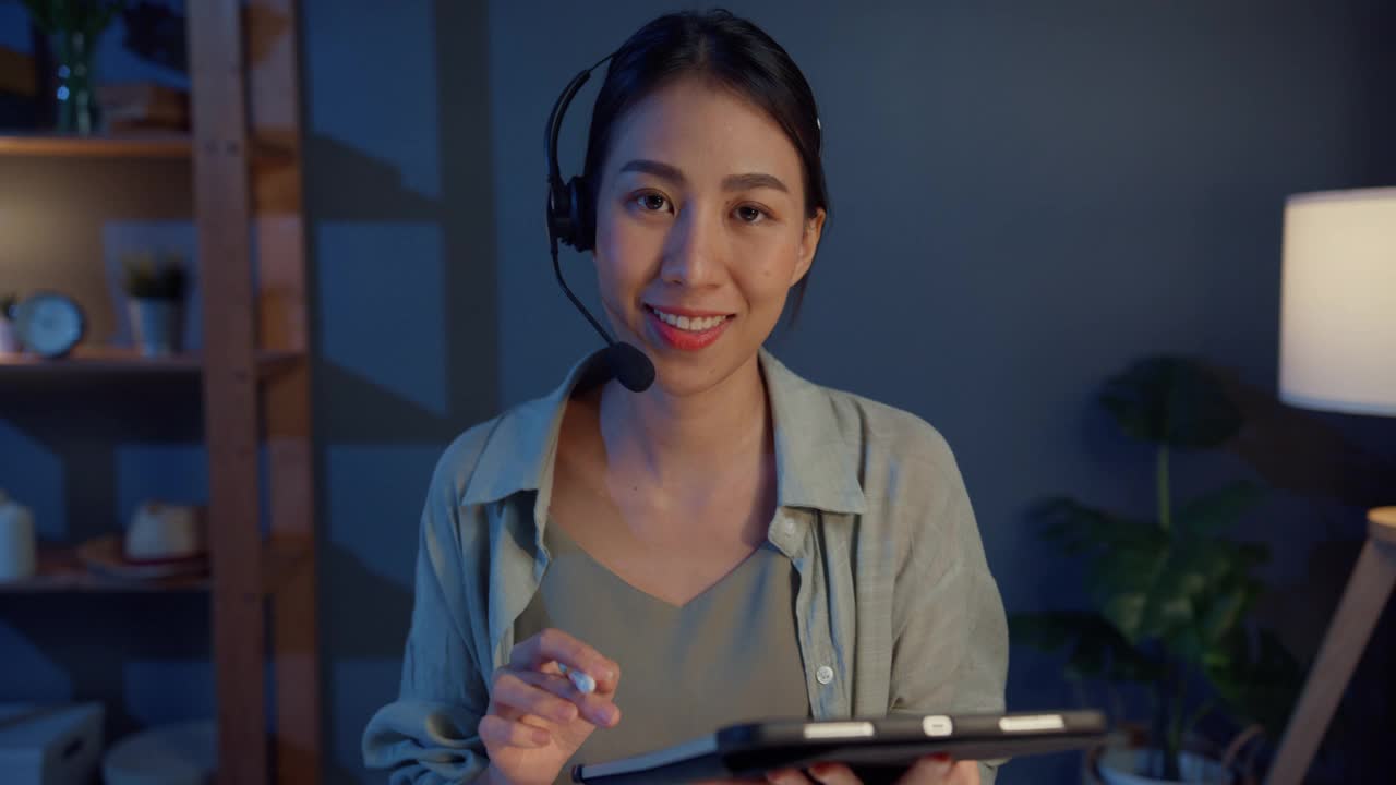 年轻的亚洲自由职业女性戴着耳机，晚上在家的客厅里用笔记本电脑进行视频会议和电话会议。视频素材