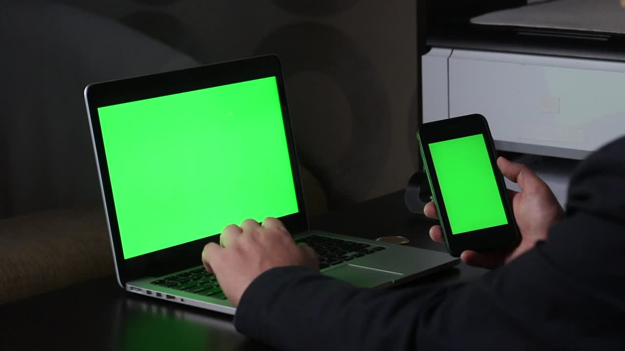 一名男子在一台绿色屏幕的笔记本电脑前工作。他手里拿着一部绿色屏幕的智能手机。附近有一枚加密货币。Chromakey视频素材