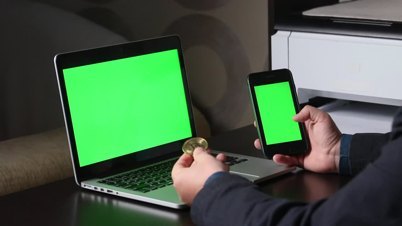一名男子在一台绿色屏幕的笔记本电脑前工作。他手里拿着一部绿色屏幕的智能手机。考虑加密货币。Chromakey。视频素材