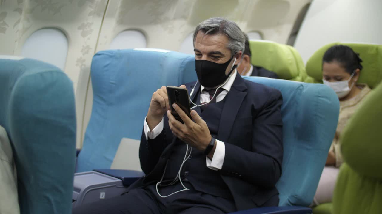 商人带着防护口罩在飞机上使用手机去国外旅行。用手机消磨无聊时间的乘客。新常态旅行。视频下载