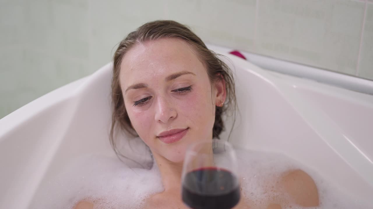 特写肖像的快乐轻松白种人年轻苗条的女人喝红酒在慢动作躺在浴缸泡沫。美丽的千禧女士享受周末在家里的浴室喝酒视频素材