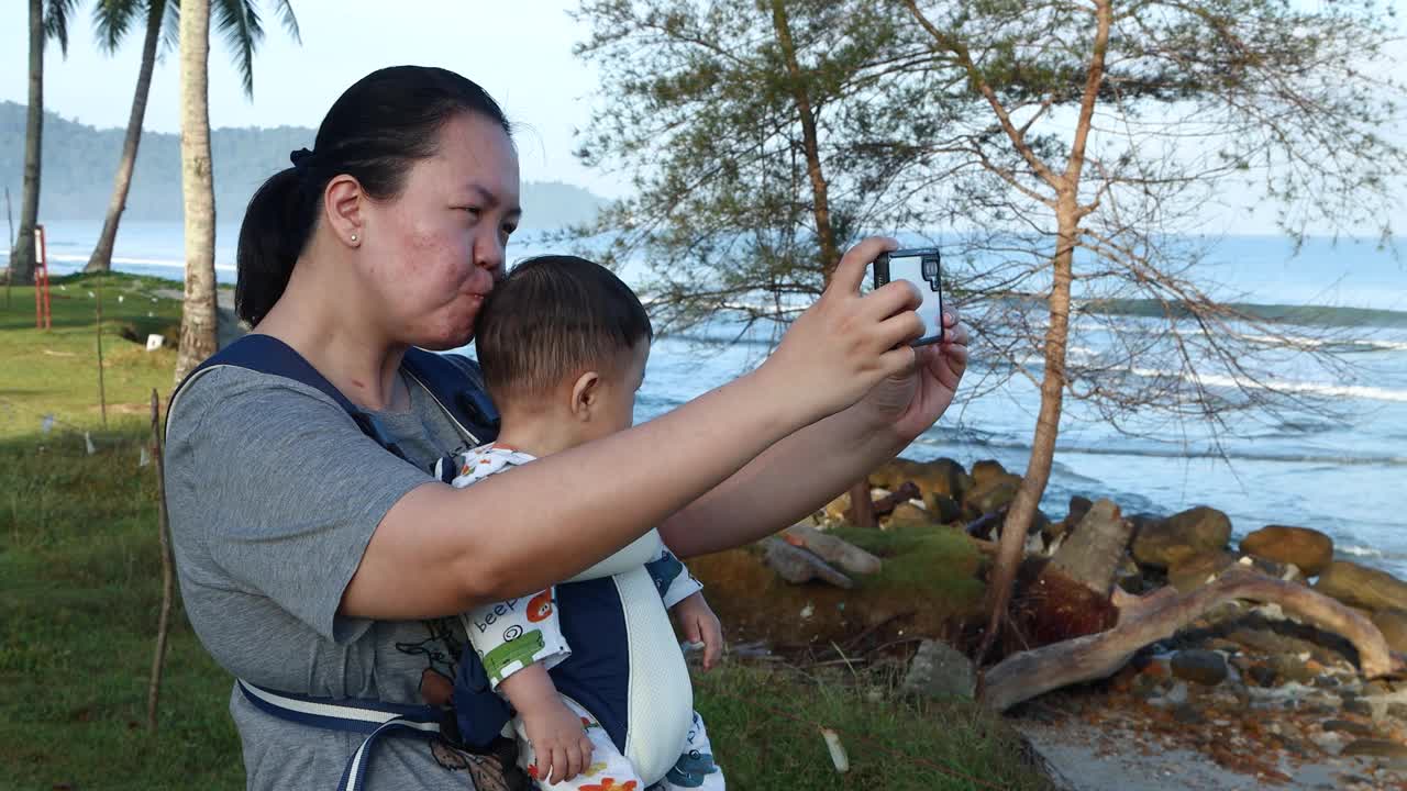 亚洲华裔母亲抱着孩子，在美丽的热带海滩上与孩子自拍。视频素材
