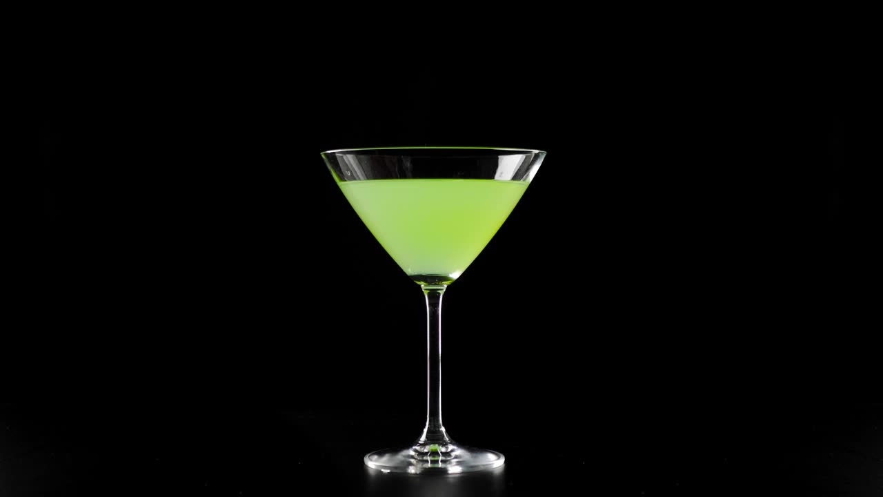 绿色鸡尾酒。黑色的桌子上，黑色的背景下，马提尼杯上的绿色夏日鸡尾酒，手将冰冷的冰块倒入杯中。酒精鸡尾酒会、夜生活和夜总会的概念。4 k视频下载