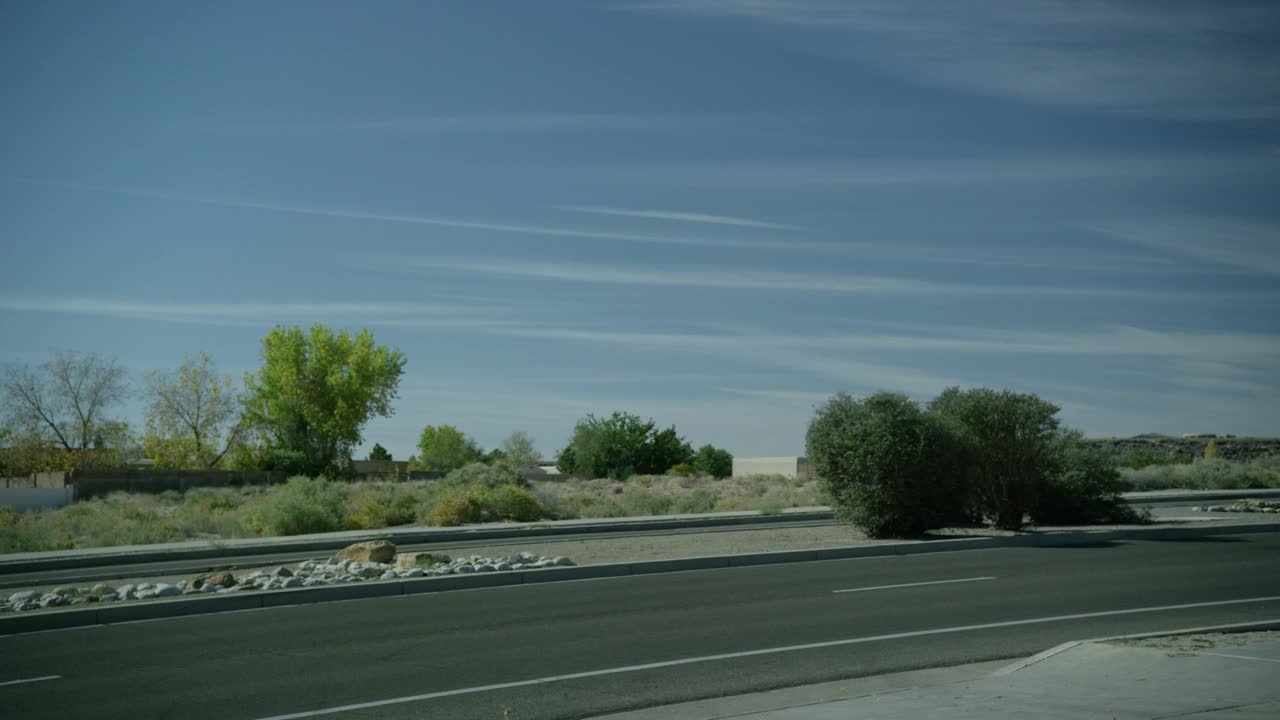 当车辆从右到左行驶时，道路的中等角度。中间有灌木和岩石可见。在bg中可见的树。农村地区。蓝色的天空。视频素材