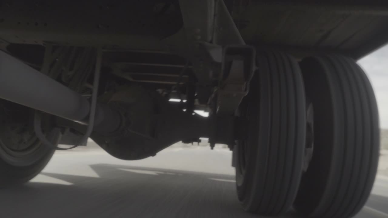 半卡车、大卡车或18轮卡车的底部或起落架的闭合角移动角度。轮胎、车轴和传动轴部分可见。卡车行驶在沙漠高速公路或高速公路上。视频素材