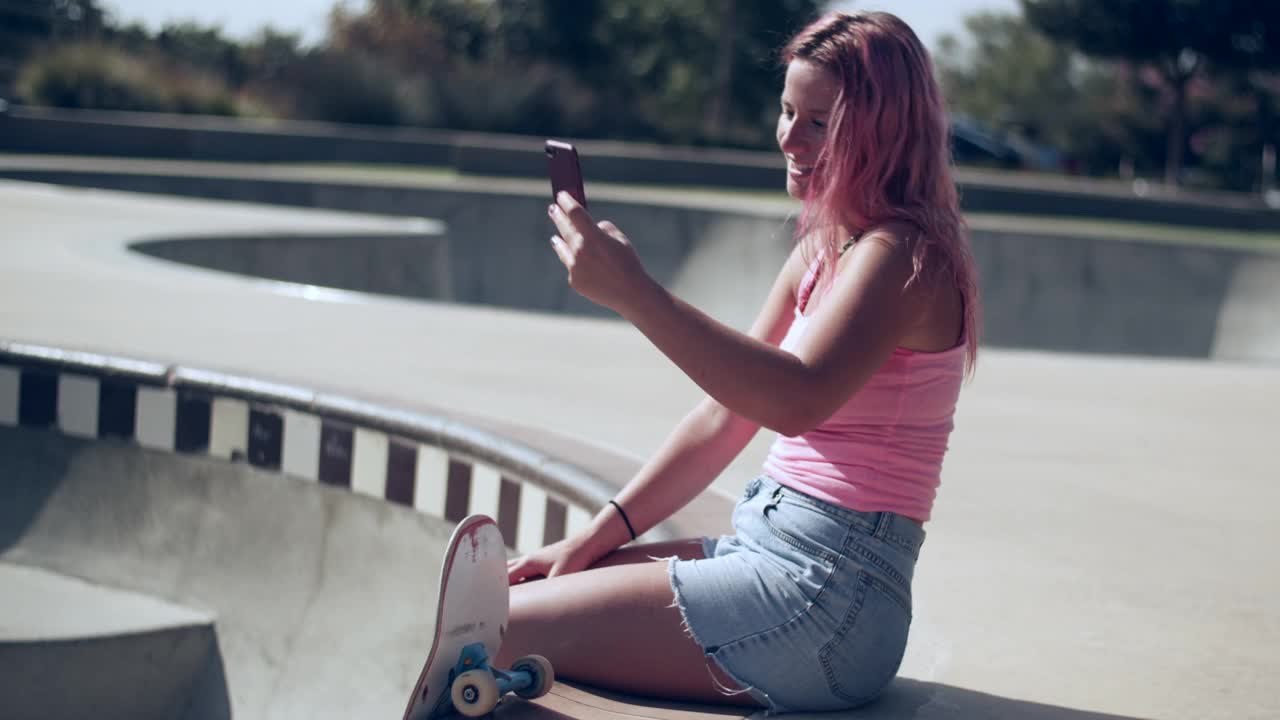 一个粉色头发的年轻女子在滑板公园玩手机视频素材