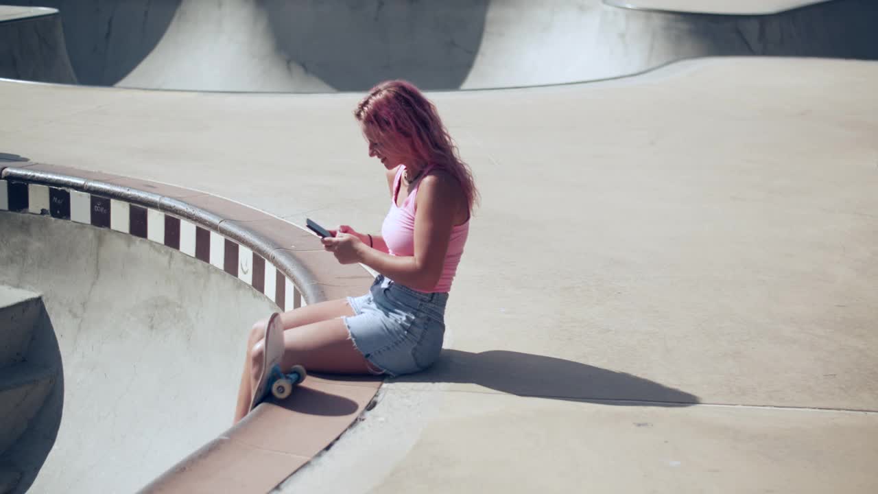 一个粉色头发的年轻女子在滑板公园玩手机视频素材