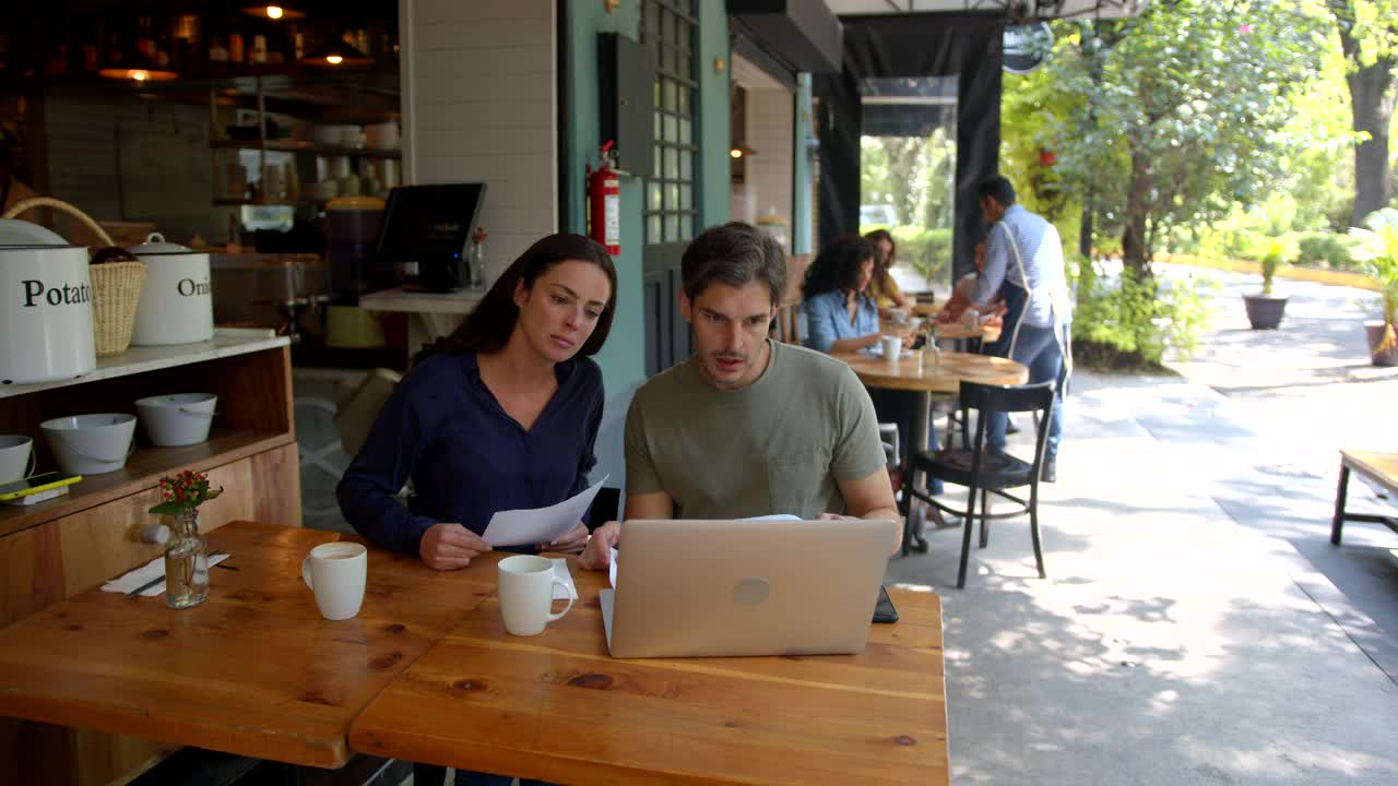 专注的商业夫妇在咖啡馆用笔记本电脑一起工作，而其他顾客在后台，服务员在为他们服务视频素材