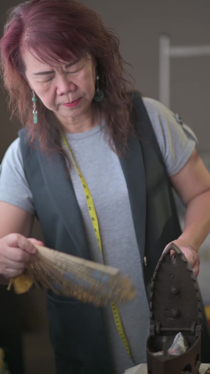 一名亚洲华裔高级女性裁缝在她的工作室里给古董铁器具扇风，在熨布之前先加热视频素材