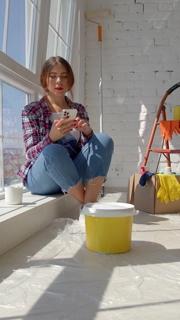 一个年轻白人女人在装修房子的视频。一位女画家拿着智能手机在地板上休息。家居概念。新屋的概念。4 k慢动作视频下载