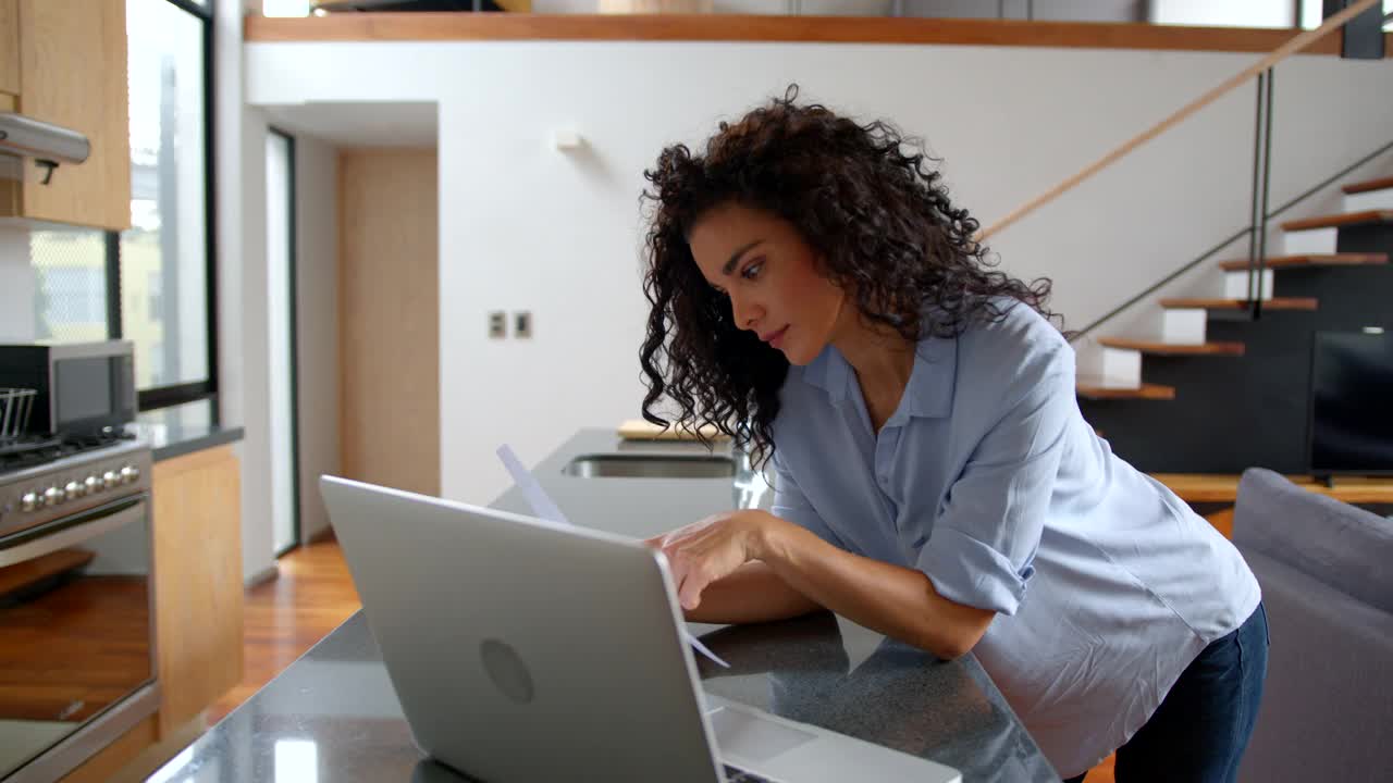 漂亮的卷发年轻女子在家里靠在厨房柜台上用她的笔记本电脑支付账单视频素材