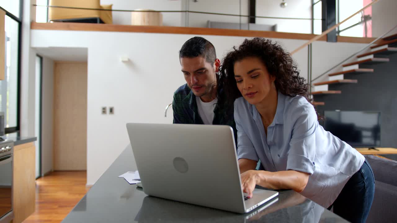 一对快乐的年轻夫妇在家里靠在厨房的柜台上看着水电费账单，一边聊天一边用笔记本电脑在线支付视频素材