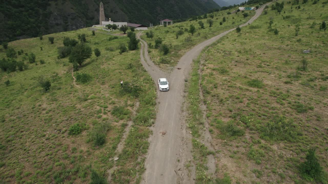 鸟瞰图的乘客白色汽车驾驶在农村狭窄的道路在山区。行动。车辆行驶在稀有的树木和绿色的山坡上。视频素材