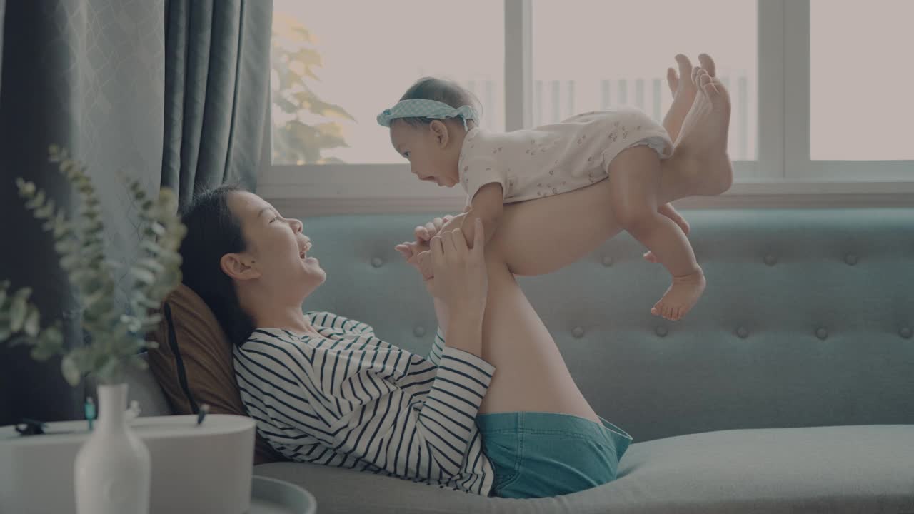 幸福团聚的亚洲妈妈躺在舒适的沙发上玩起与女儿(5-8个月)的家庭生活视频素材