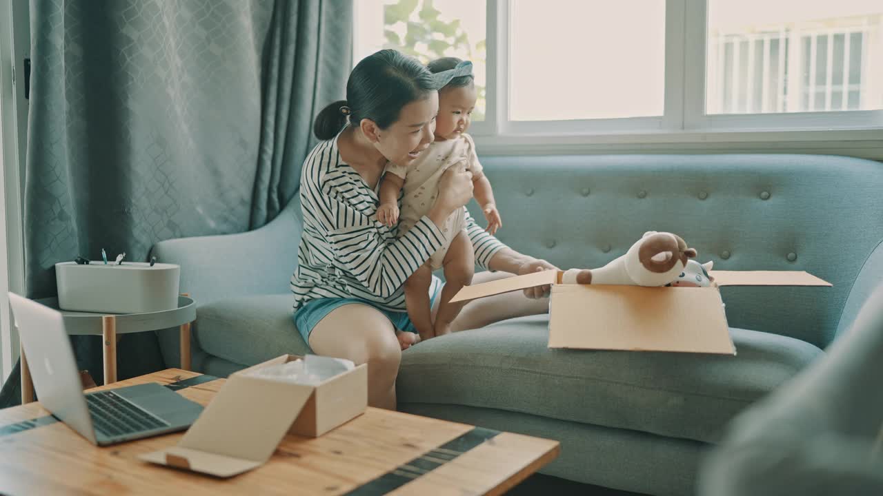 亚洲年轻的成年母亲在舒适的沙发上为女儿(5-8个月)购买网上购物玩具和打开盒子的家庭生活视频素材