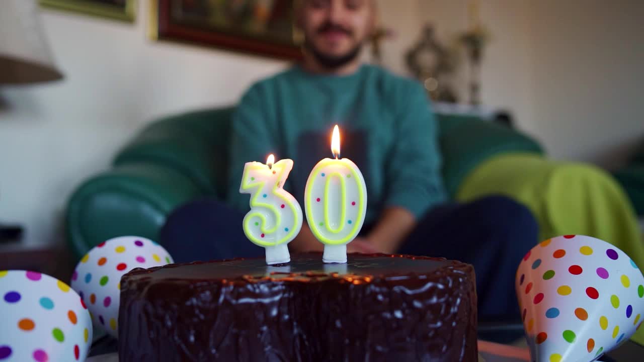 兴奋的年轻人吹着生日蛋糕上的蜡烛，在家里庆祝他的30岁生日视频下载