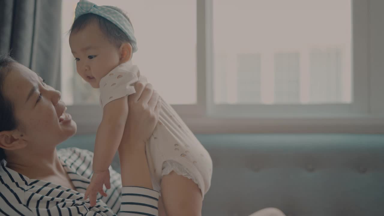 亚洲年轻的成年母亲在舒适的沙发上玩耍和亲吻女儿(5-8个月)的家庭生活视频素材