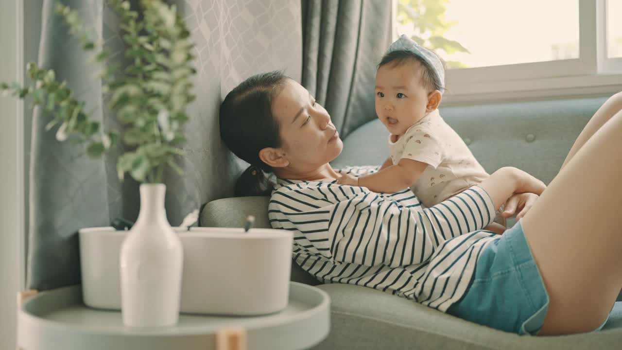 幸福团聚的亚洲妈妈躺在舒适的沙发上玩起与女儿(5-8个月)的家庭生活视频素材