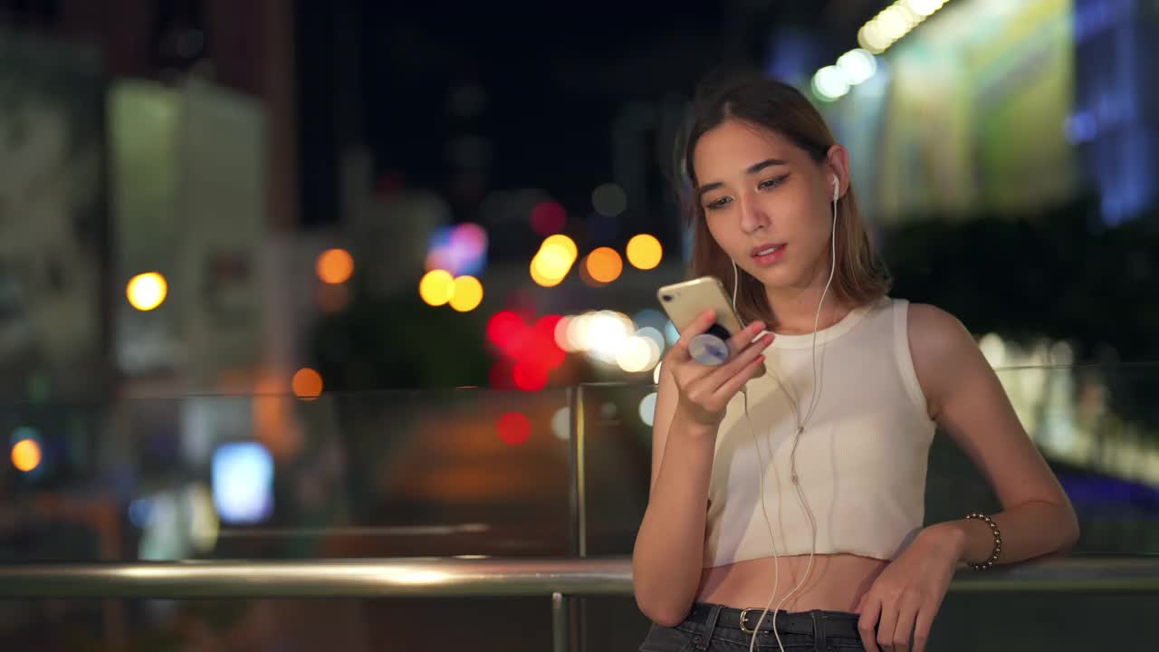 年轻亚洲女人的4K肖像走在城市的街道听音乐从耳机在晚上视频素材
