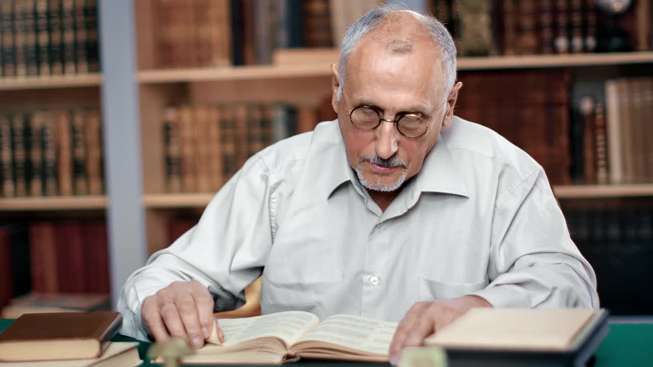 老年男子专业历史教授坐在图书馆办公桌旁学习古玩书视频素材