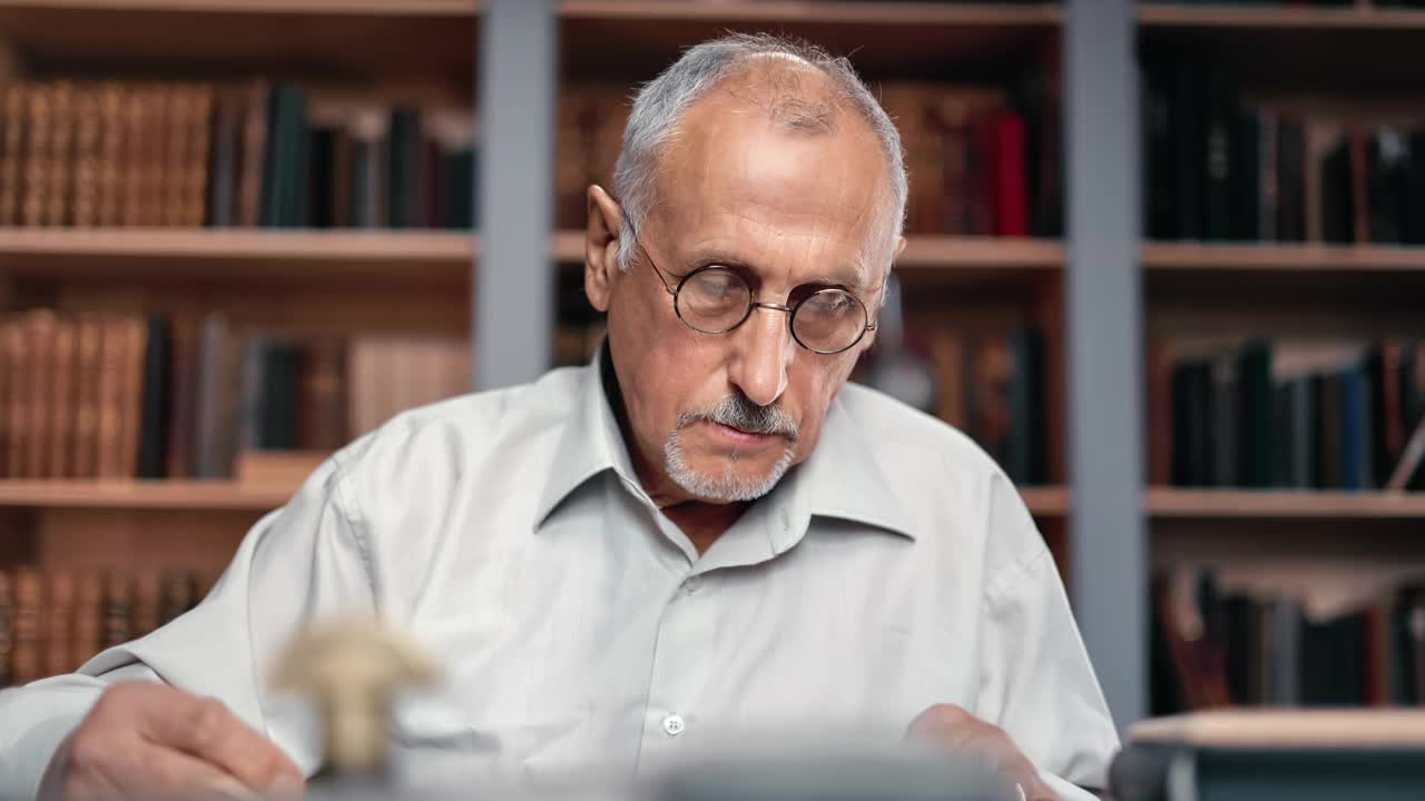 特写一位白发苍苍的老人在图书馆书桌前阅读书籍，搜寻历史研究视频素材
