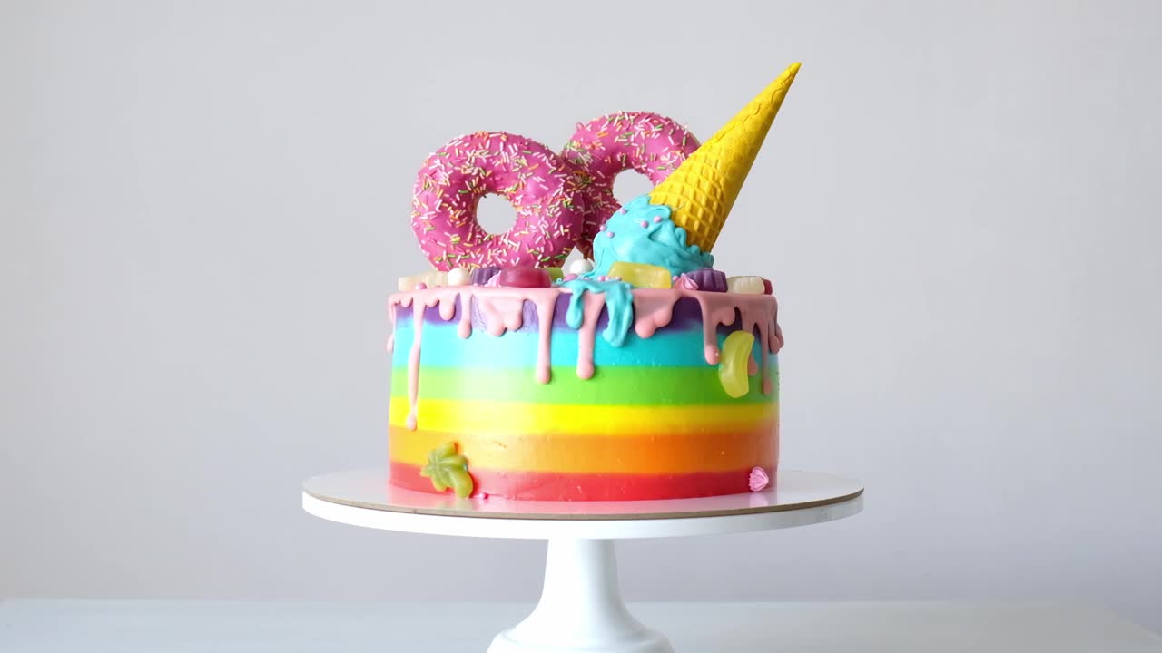 生日蛋糕，白色背景上有五彩缤纷的彩虹奶油，装饰有华夫饼蛋筒，粉色甜甜圈，糖屑，橘子酱糖果浇满巧克力。视频下载