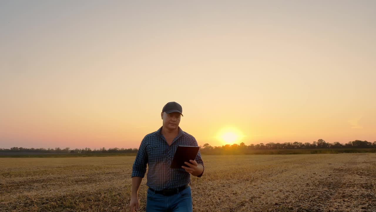 农夫在日落时散步。农业。农民的剪影。法默手里拿着数码平板电脑，在夕阳西下或旭日升起的时候走过一片被割过的田地。农业经营理念视频素材