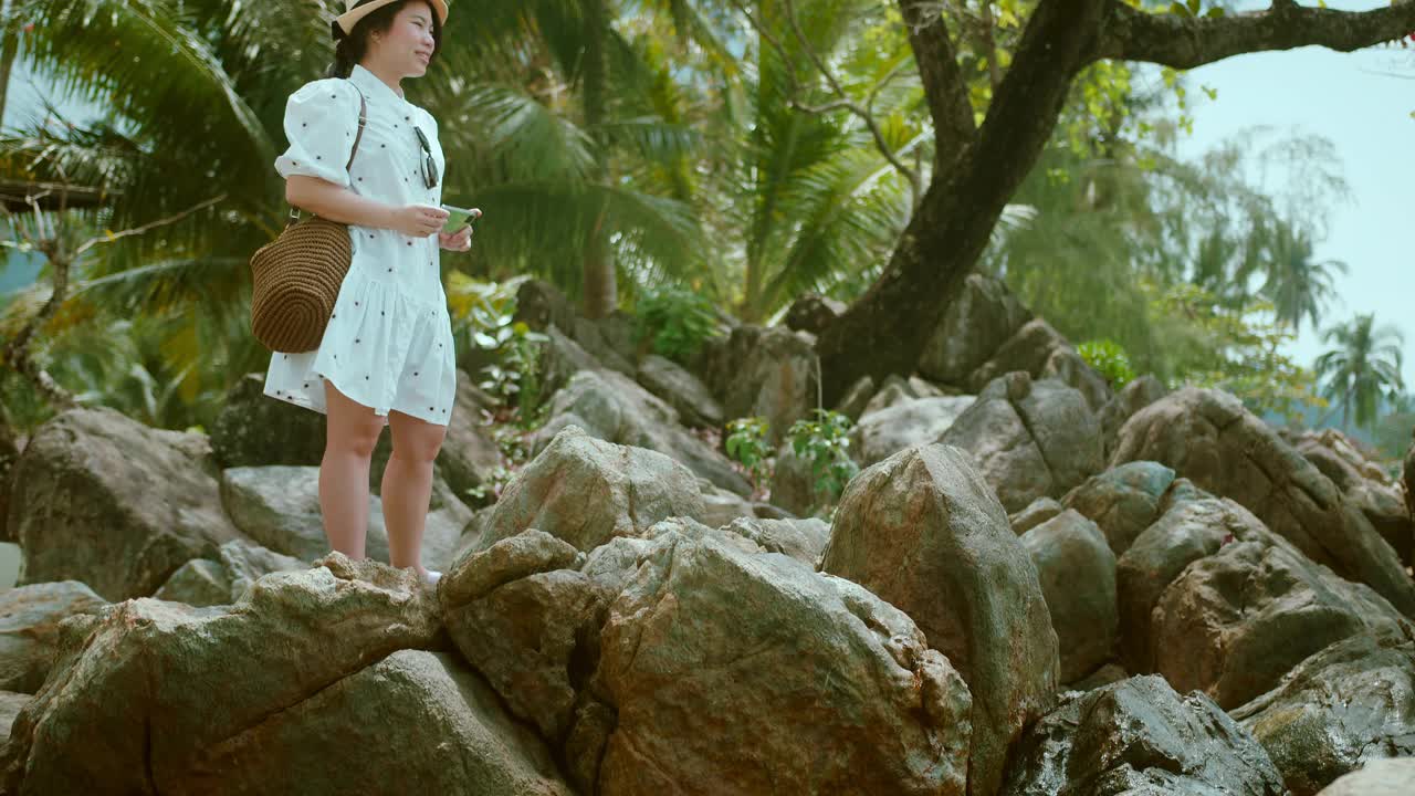 迷人的亚洲女性旅行者穿着休闲布夏季周末假期手握智能手机视频通话社交媒体与朋友的快乐和幸福在海滩海洋自然背景视频下载