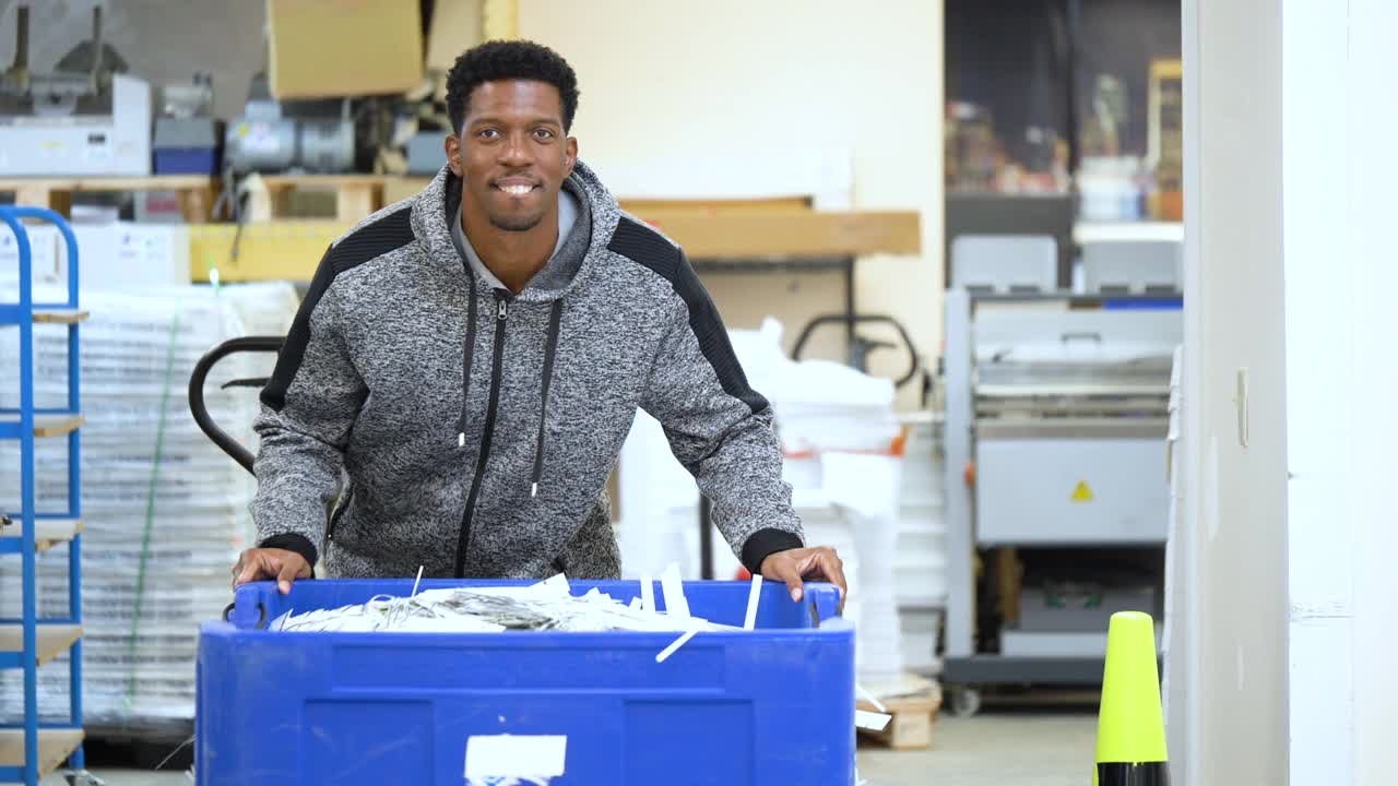 一个黑人在印刷厂推着一个巨大的回收箱视频素材