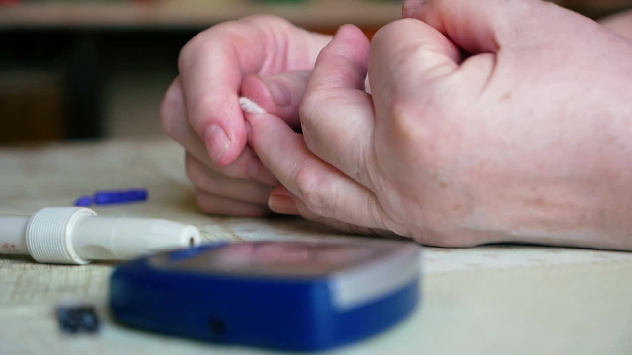 一个糖尿病患者在家里用血糖仪验血后的手特写。表上的表焦不清。这位妇女用棉签止住了手指上的血。视频下载