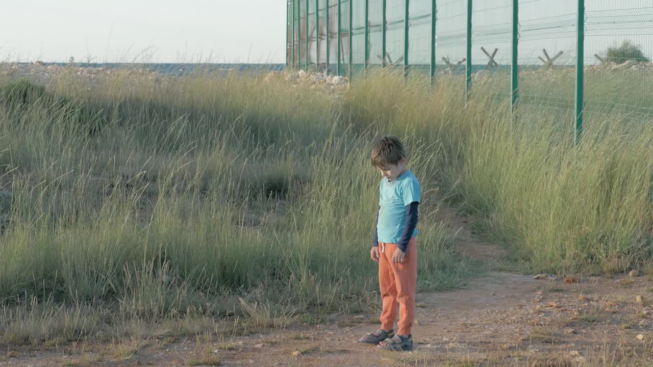 一个穿着脏衣服的小男孩孤独地站在金属栅栏附近的田野里看着障碍物。概念感受的情绪难民的悲伤和抑郁视频下载