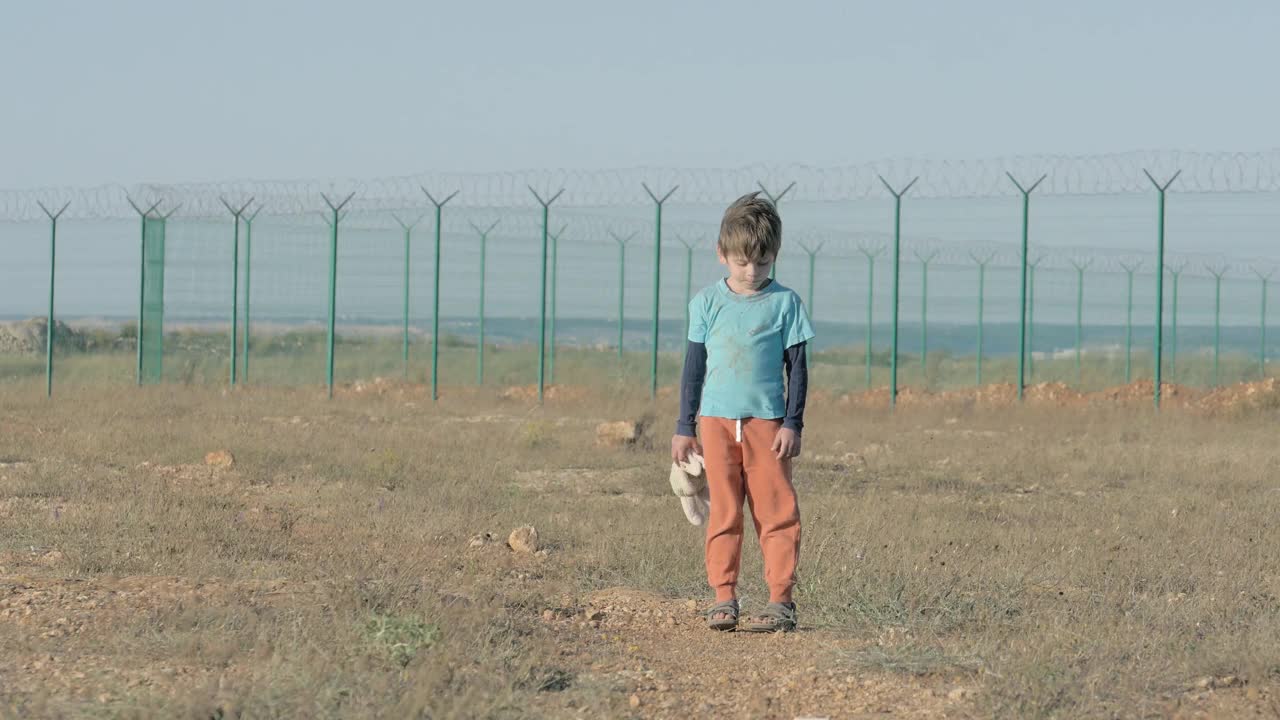 伤心的小男孩孤儿走在废弃的路上，手里拿着一只玩具兔子，低着头，停下来望着远方。移民和难民营的概念视频下载