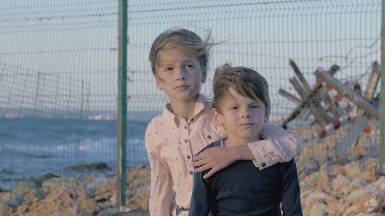 抑郁的孩子。孤独的女孩在伤心的男孩身边哭泣。悲伤的小难民兄弟姐妹在边境的岸边。沿海有带刺铁丝网的保护区视频下载