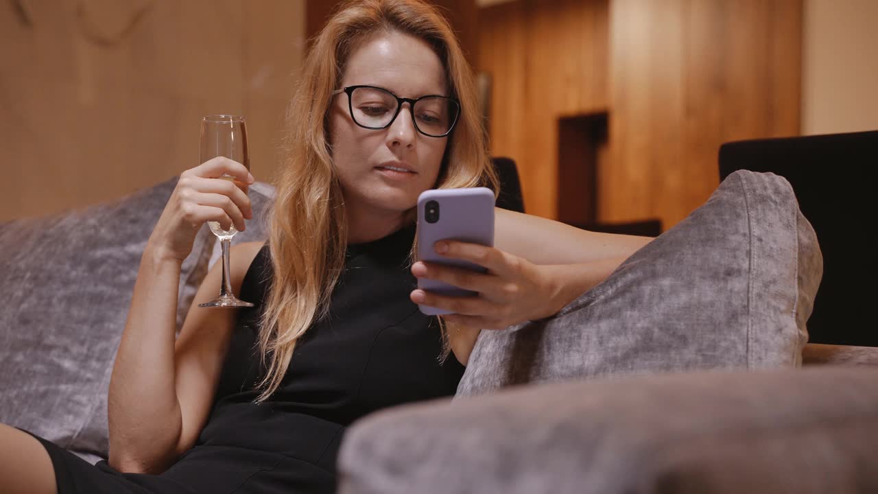 穿着晚礼服的女人拿着酒杯拿着电话坐在沙发上大笑视频素材