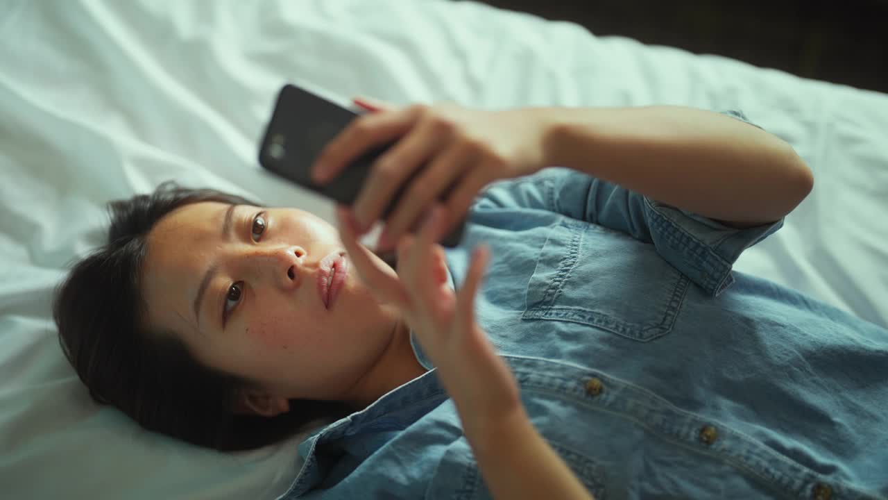年轻女子躺在床上使用智能手机的高角度照片视频素材