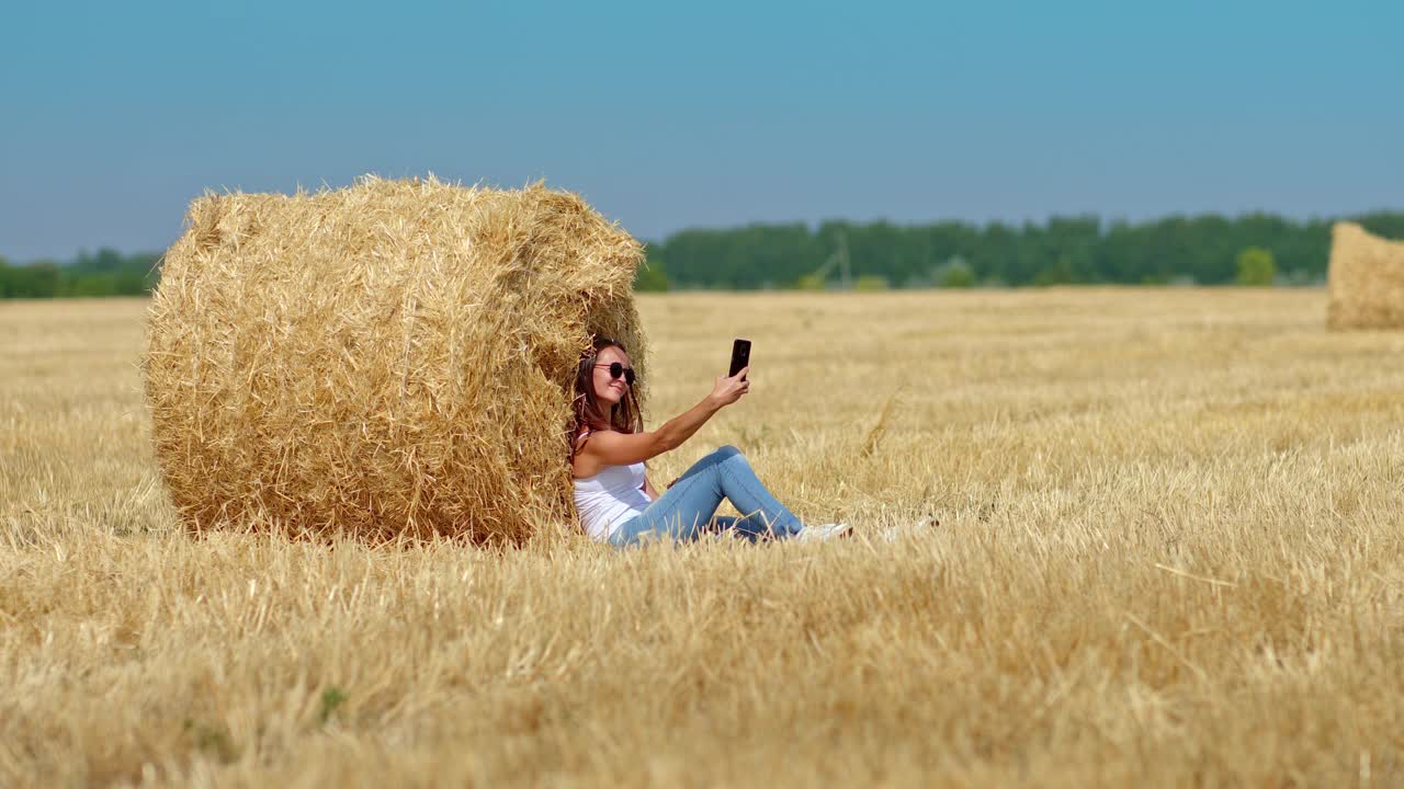 一个善良的女人在麦田里自拍。一名女子坐在干草堆旁，微笑着用智能手机拍了张照片。她是快乐的。4 k, ProRes视频下载