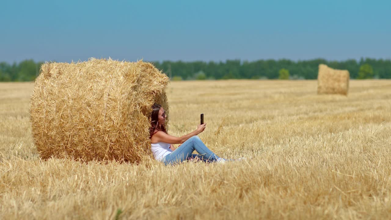 年轻女子在麦田里自拍，她坐在一个大干草堆旁。美丽的女人正微笑着用手机拍照。田园风光，麦田里的干草堆。4 k, ProRes视频下载