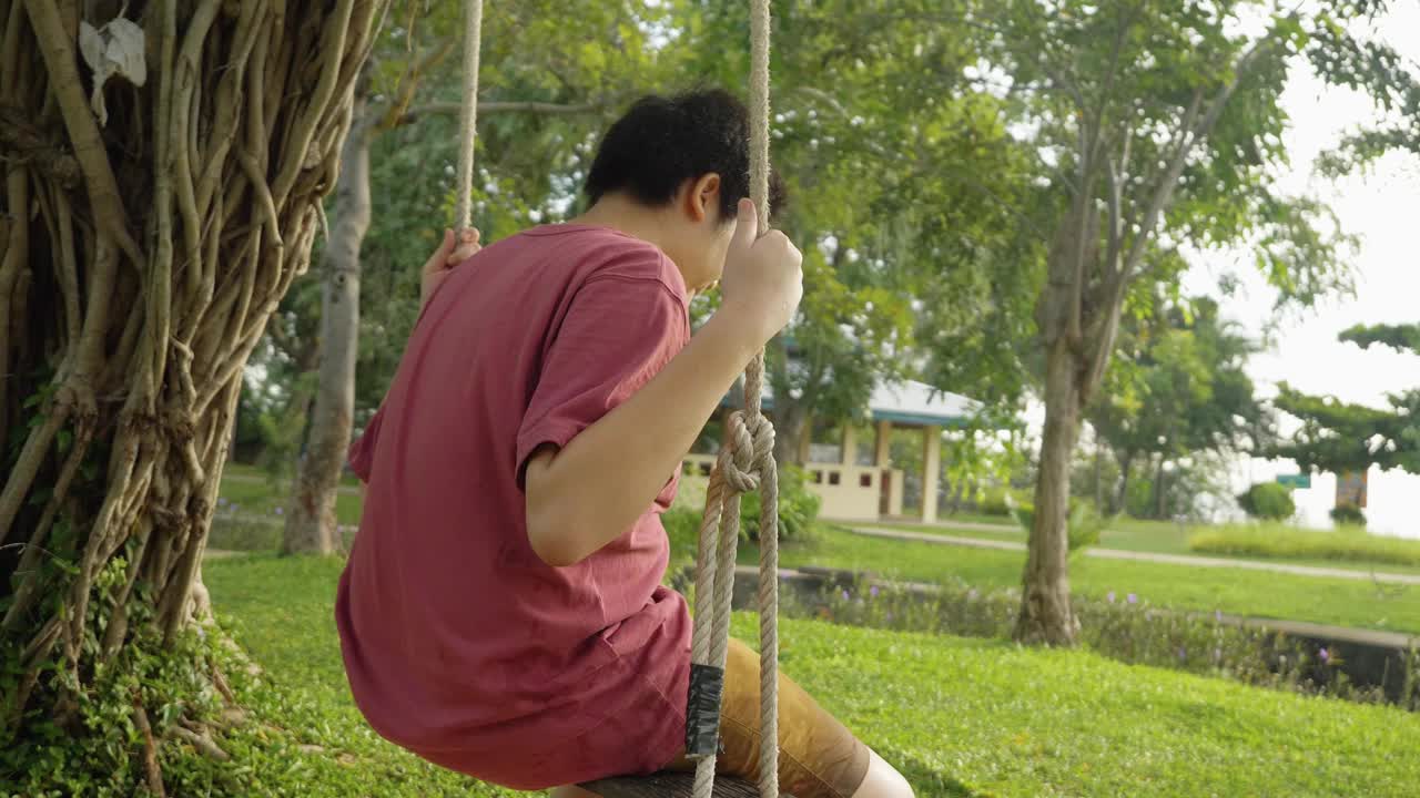 亚洲男孩坐在户外公园树下悬挂的长袍秋千上，生活理念。视频素材